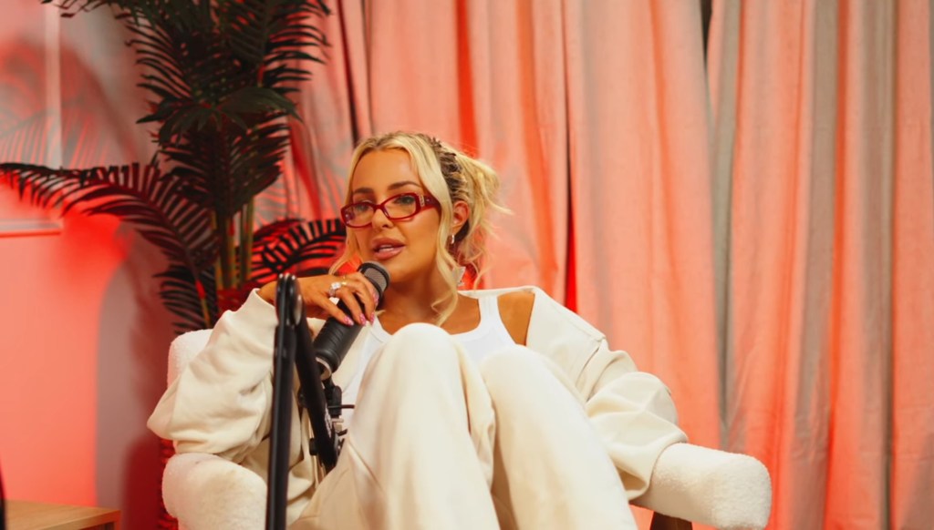 Tana Mongeau wearing red glasses and a white jumpsuit, sitting in a white chair holding a microphone on the 'Cancelled' podcast set, with an orange illuminated curtain backdrop.