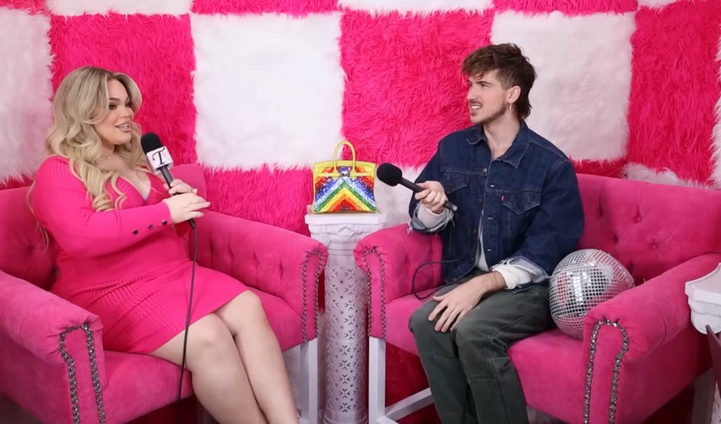 Trisha Paytas talking to Joey Graceffa on the 'Just Trish' podcast set, with a rainbow Birkin bag placed on a white pedestal between two bright pink chairs where Trisha and Joey are seated.