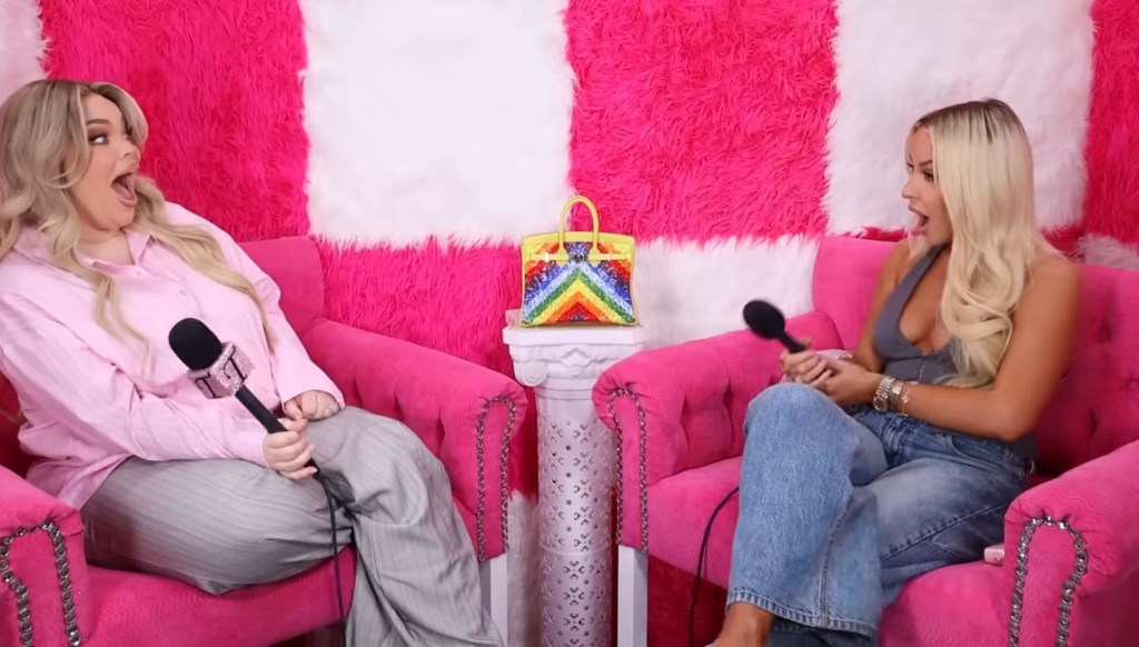 Tana Mongeau and Trisha Paytas on the 'Just Trish' podcast set, both with their mouths wide open in shock, sitting in bright pink chairs with a rainbow Birkin bag on a white pedestal between them.