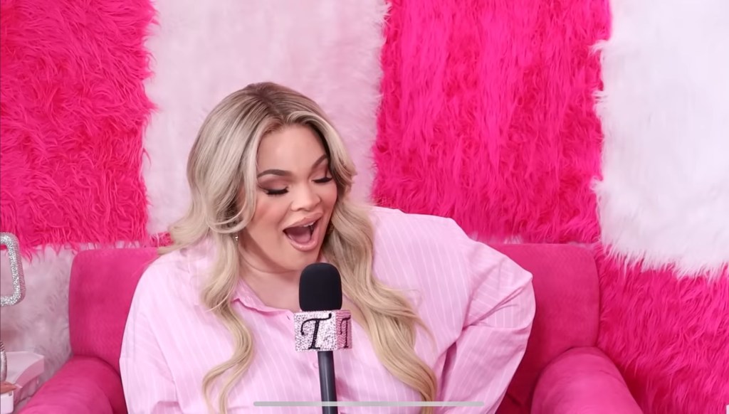 Medium close-up shot of Trisha Paytas on the 'Just Trish' podcast, with her mouth wide open and eyes closed in shock, set against a plaid bright pink and white background.
