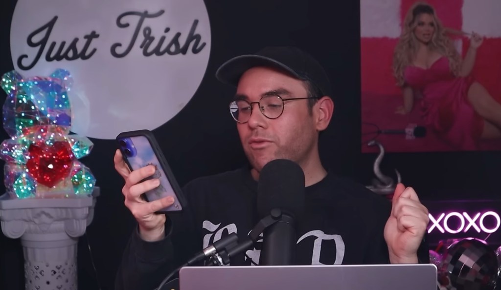 Oscar Gracey filming Just Trish talking into his microphone whil holding his phone. There's a crystal bear on his left and a post of Trisha Paytas on his right in the background. 