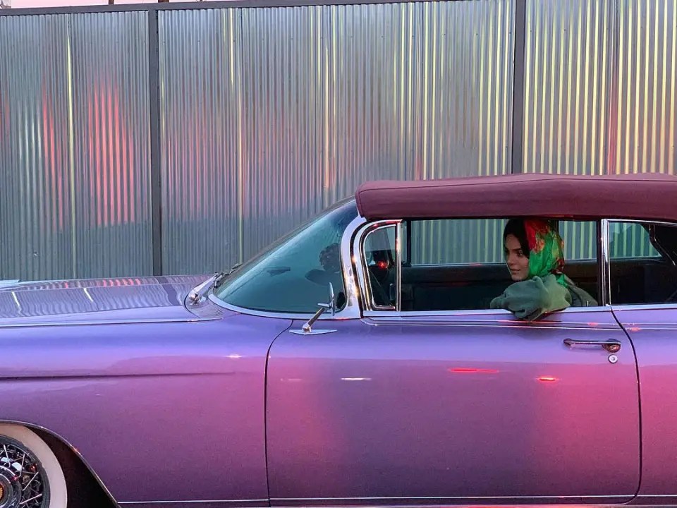 Image of Kendall jenner sitting inside her vintage, hot pink 1960 cadillac in fron of a silver background. She is wearing a green and red head scarf with her arm rested on the window sill and a slight smirk. 