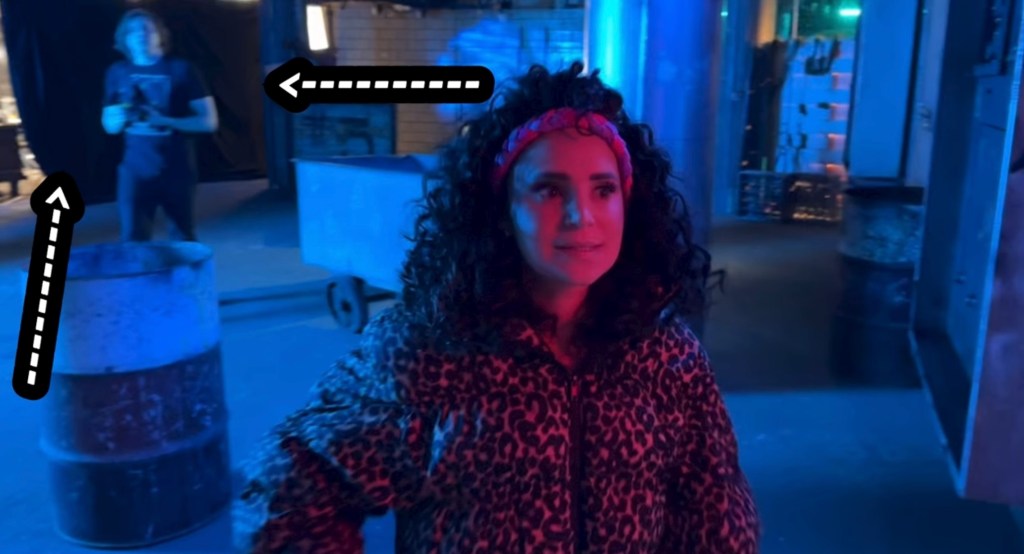 Behind the scenes of Escape the Night Season 5. Rosanna Pansino stands in the center of the screen with curly hair with two arrows pointing to a black curtain in the top left corner of the picture.