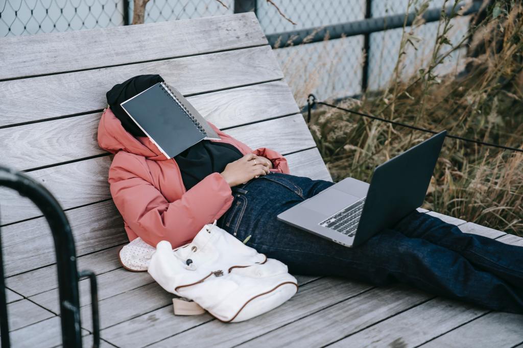 person laying on wooden platform with laptop on legs and notebook open covering face
